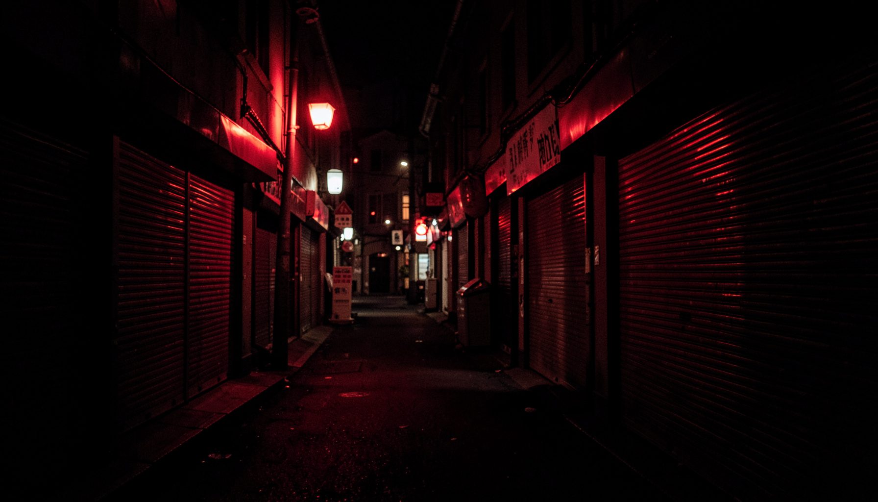 夜の商店街のシャッターが並ぶ通り。赤い街灯が点々と灯り、深い影の中に温かな光が浮かぶ、もの悲しい雰囲気の写真